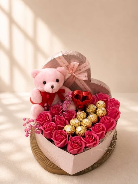 Heart-shaped gift box with pink roses, chocolates, red heart decoration, and pink teddy bear with bow on beige background