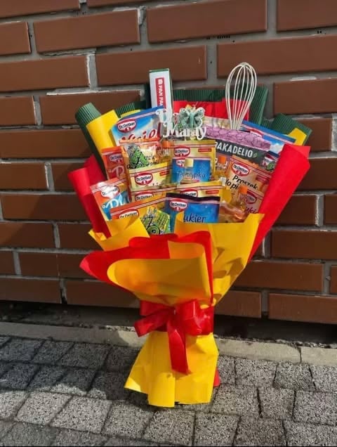 Decorative bouquet made of packaged foods and kitchen utensils wrapped in yellow and red paper with a red bow against a brick wall