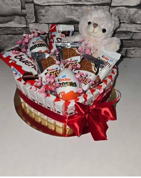 Heart-shaped gift basket filled with Kinder chocolates, candies, and pink flowers, topped with a white fluffy teddy bear and red ribbon bow