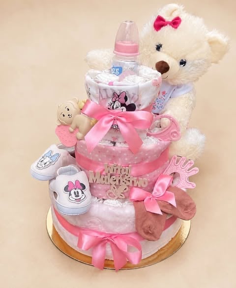Three-tiered pink diaper cake decorated with a white stuffed animal, baby bottles, and pink ribbon bows on a gold base