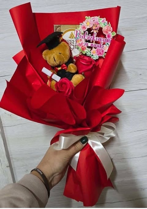 Hand holding a red wrapped bouquet with pink flowers, teddy bear, and chocolates on white wooden background