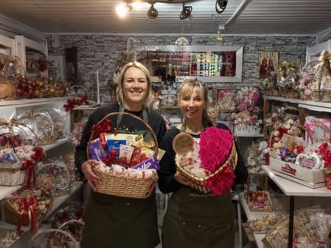 Two women smiling in a gift shop holding heart-shaped baskets with chocolates and gifts, surrounded by holiday decorations and merchandise on shelves