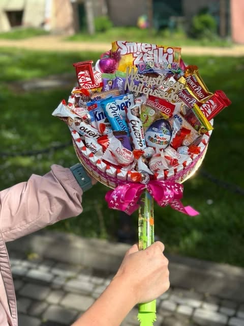 Hand holding a colorful candy bouquet made of assorted snacks and sweets with a pink bow, in a garden setting